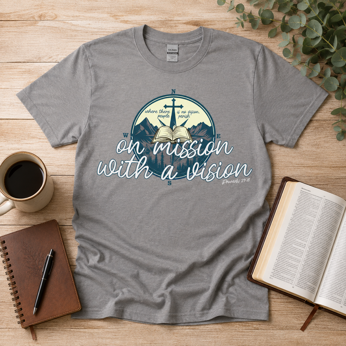 A heather gray graphic t-shirt laid flat on a wooden desk with a coffee mug, notebook, and open book around it. The t-shirt features a circular graphic with mountains, a cross, an open book, and the text 'on mission with a vision' and 'Exodus 34:28'.