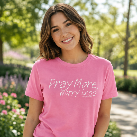 A smiling woman wearing a pink crewneck t-shirt with the white printed text 'Pray More, Worry Less' and 'Philippians 4:13' on the front, set against a blurred green park background.
