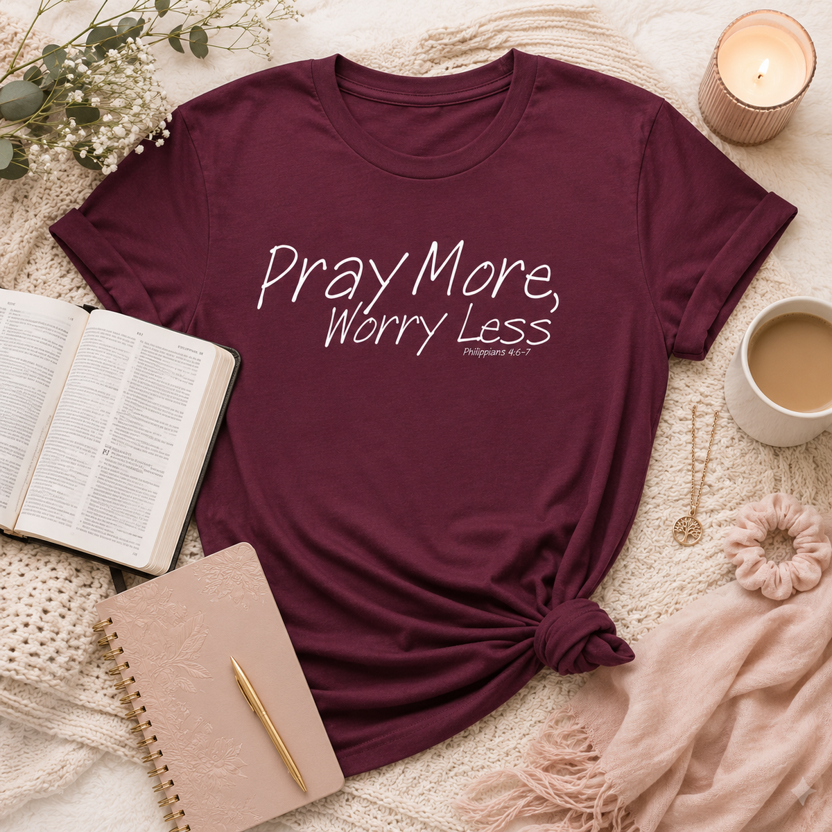 A maroon short-sleeve graphic T-shirt laid flat on a cozy beige knit blanket with a knotted hem, surrounded by lifestyle props including an open book, notebook, candle, mug of coffee, and jewelry. The shirt has white text reading 'Pray More, Worry Less' with 'Philippians 4:6-7' underneath.