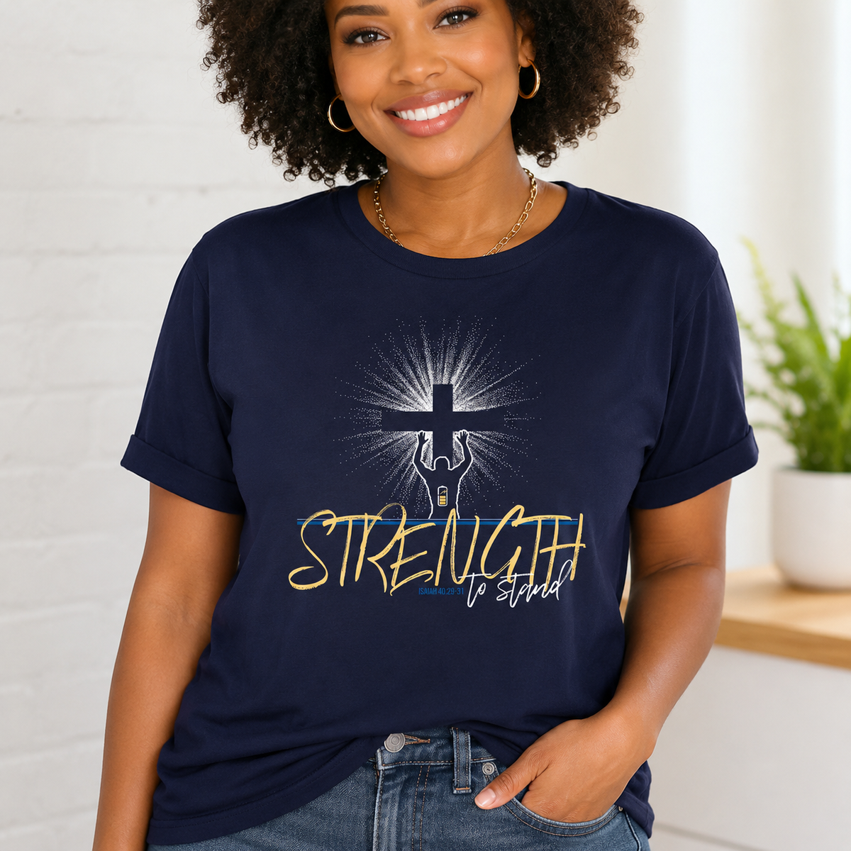 A smiling woman wearing a navy blue graphic t-shirt with a cross and the words 'STRENGTH to stand' printed on the front, paired with blue jeans in a bright indoor setting.
