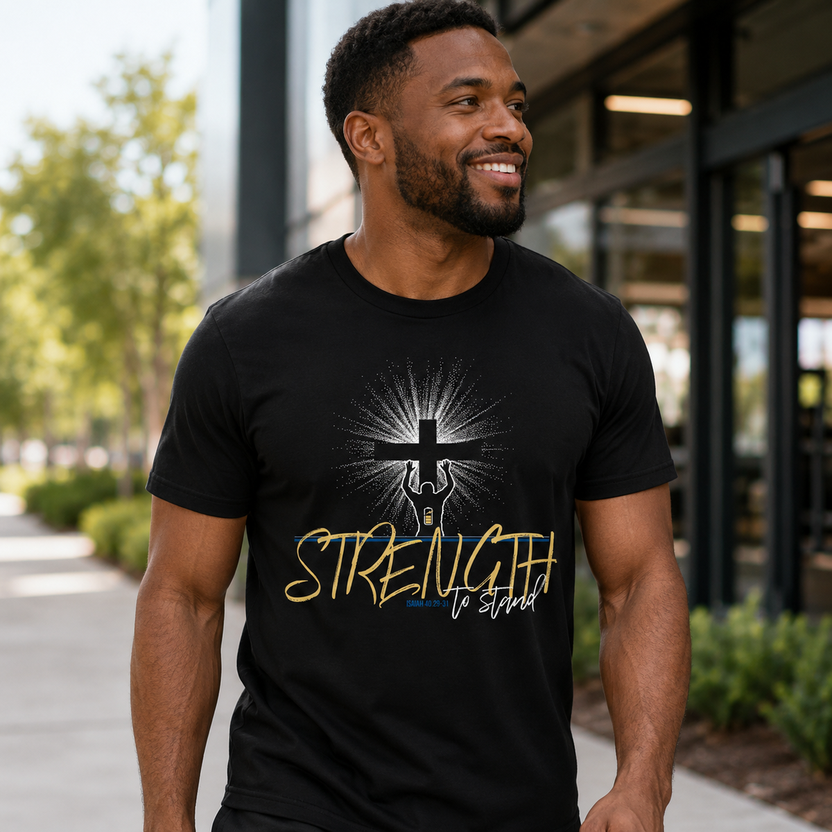 A man wearing a black graphic t-shirt with a cross and the words 'STRENGTH to stand' in gold and white, standing outdoors in front of a modern building.