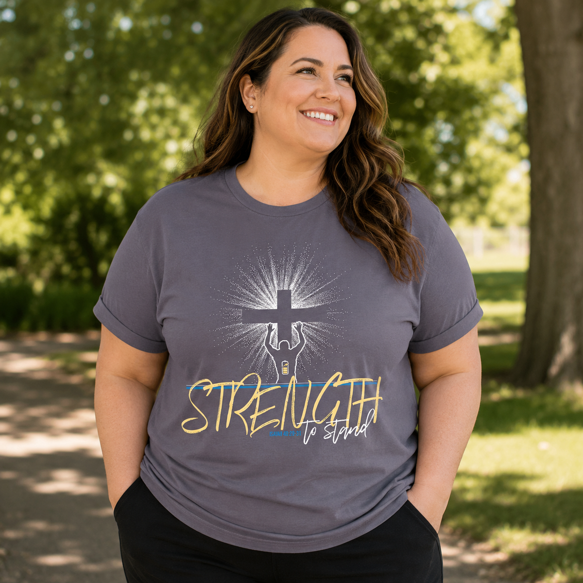 A woman wearing a heather gray graphic t-shirt with a cross and the words 'STRENGTH to stand' printed on the front, standing outdoors in a park with trees in the background.