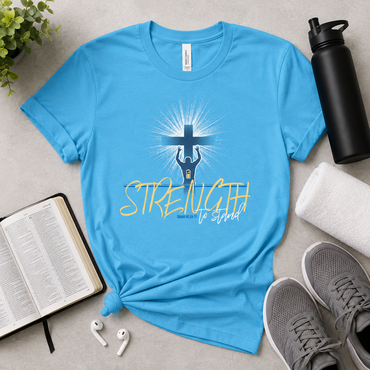 A bright blue graphic T-shirt laid flat on a light gray surface, surrounded by lifestyle props including gray sneakers, a black water bottle, a rolled towel, an open book, white earbuds, and a small plant. The shirt has a central graphic of a hand holding a cross with rays of light and the words 'STRENGTH to Stand' in yellow and dark blue.