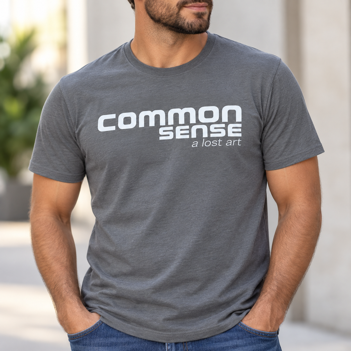 A man wearing a heather gray crewneck t-shirt with the text 'common SENSE a lost art' printed in white across the chest, paired with blue jeans.
