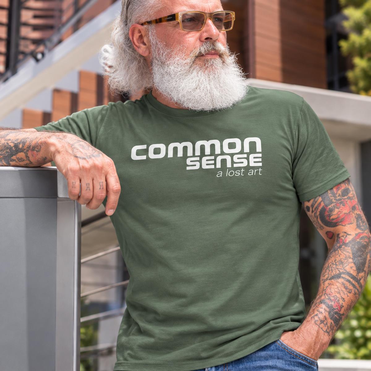 A man with a white beard and glasses wearing an olive green t-shirt with the text 'common sense a lost art' printed in white on the chest, leaning on a modern outdoor staircase.
