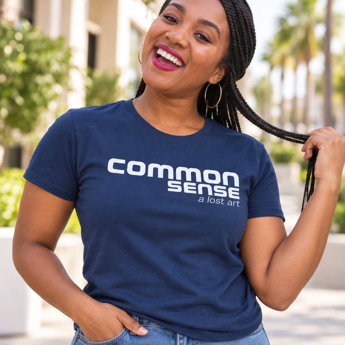 A woman wearing a navy blue graphic t-shirt with the text 'common sense a lost art' in white, posing outdoors in front of a blurred building and palm trees.