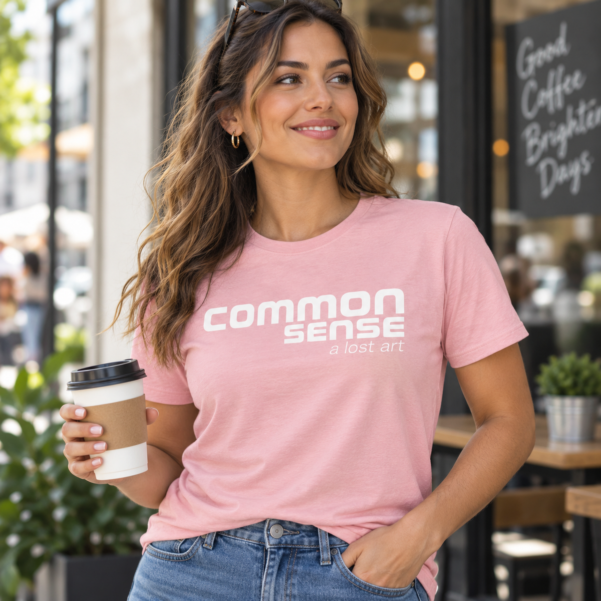 A woman wearing a light pink graphic t-shirt with the text 'common sense a lost art' in white, holding a coffee cup in front of a cafe storefront.