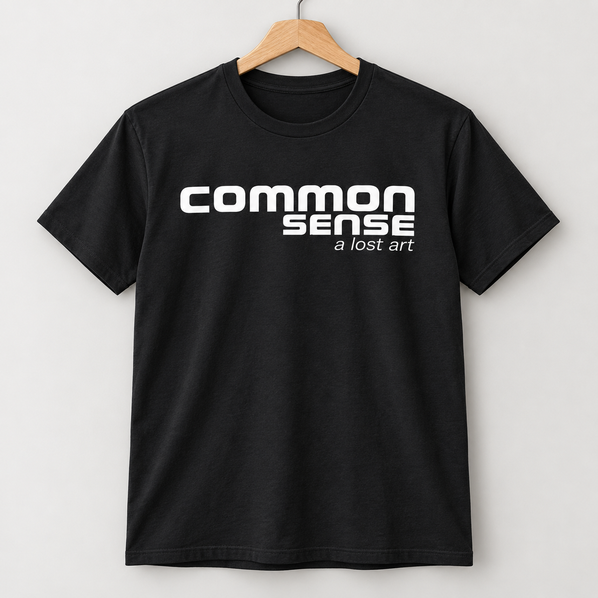 A black short-sleeve crewneck t-shirt hanging on a wooden hanger against a light gray background. The shirt features the text 'common sense' in a bold white sans-serif font, with 'a lost art' printed underneath in smaller white text.