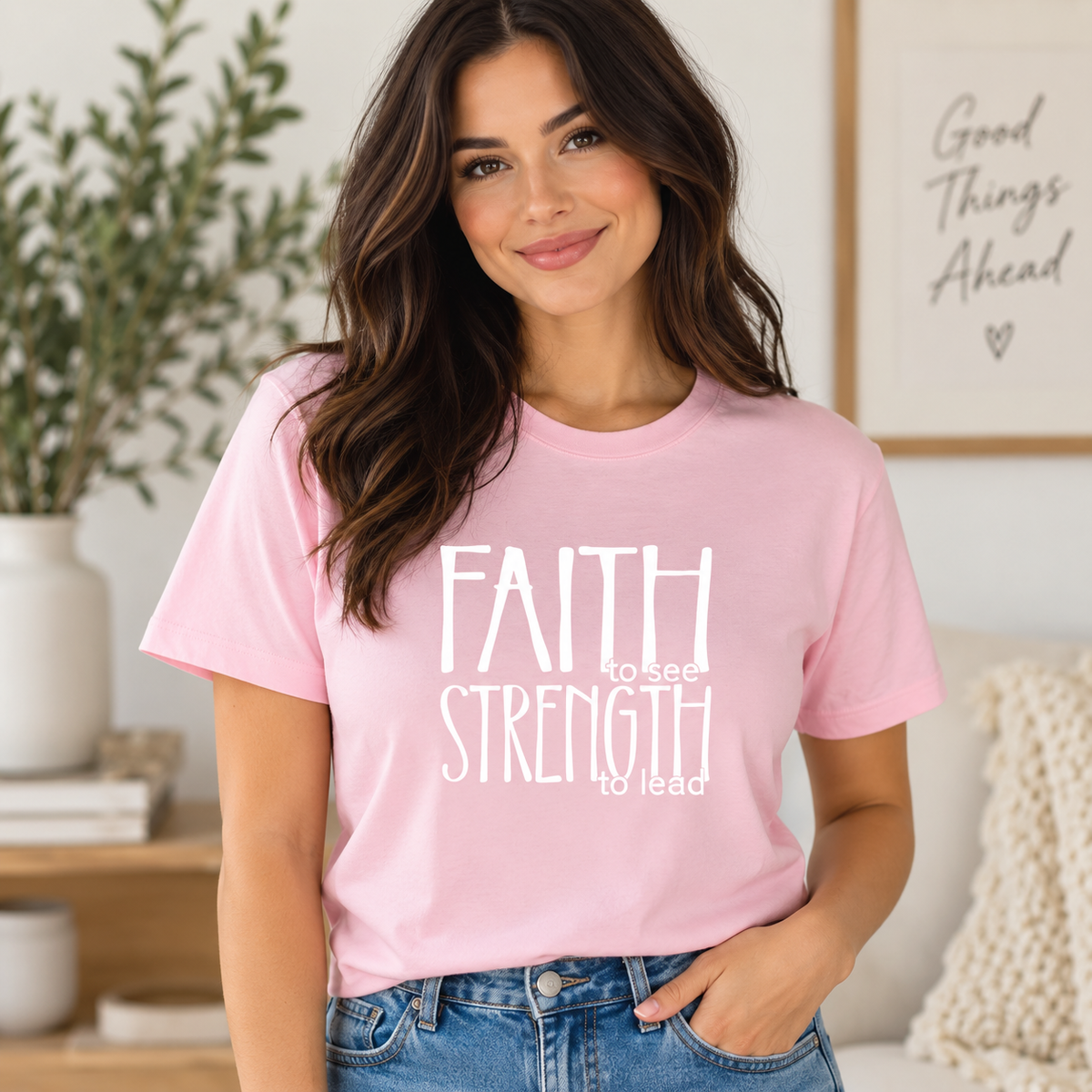 A woman wearing a light pink graphic t-shirt with the white text 'FAITH to see STRENGTH to lead' printed on the front, paired with blue jeans in a cozy, sunlit room.