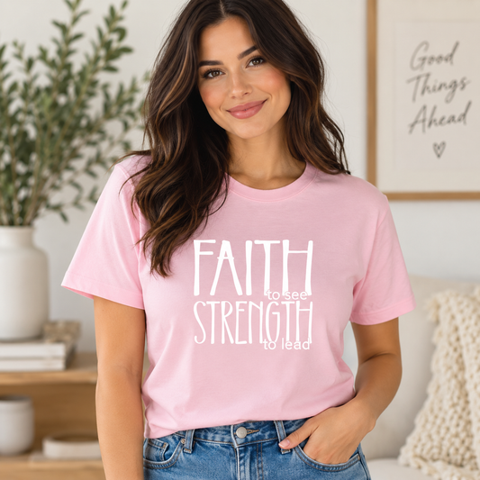 A woman wearing a light pink graphic t-shirt with the white text 'FAITH to see STRENGTH to lead' printed on the front, paired with blue jeans in a cozy, sunlit room.