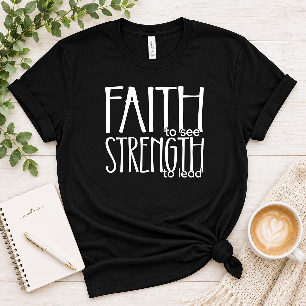 A black short-sleeve graphic T-shirt laid flat on a light wood surface, styled with a knot at the bottom hem. The shirt has large white text reading “FAITH to see STRENGTH to lead.