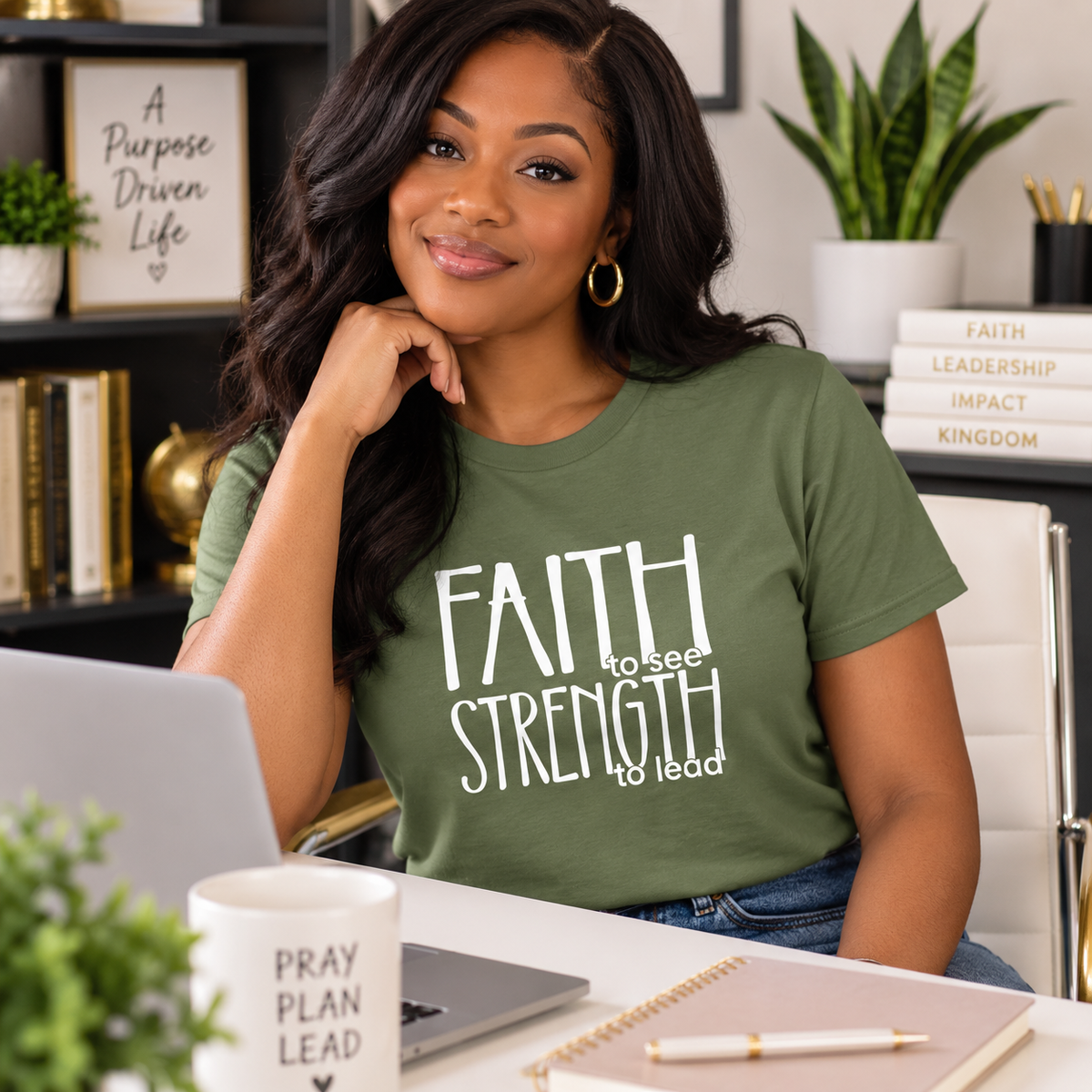 A woman with long dark hair sits at a desk wearing an olive green graphic t-shirt with the white text 'FAITH to see STRENGTH to lead'. The setting is a bright, modern home office with a laptop, notebook, and plants in the background.