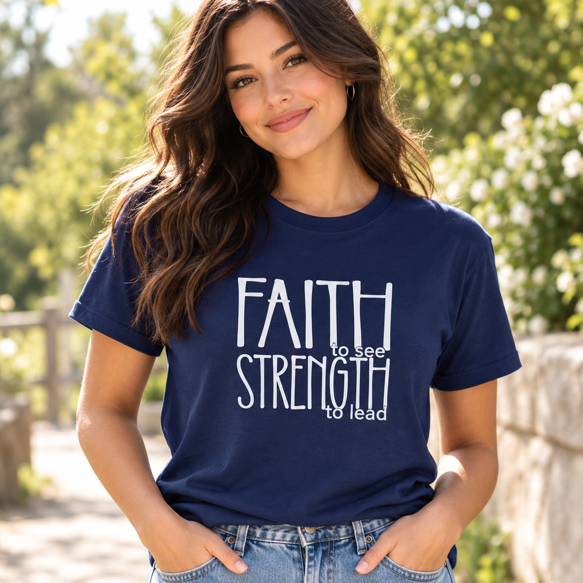 A woman wearing a navy blue graphic t-shirt with the text 'FAITH to see STRENGTH to lead' in white, paired with blue jeans, set against a blurred outdoor background of green foliage.