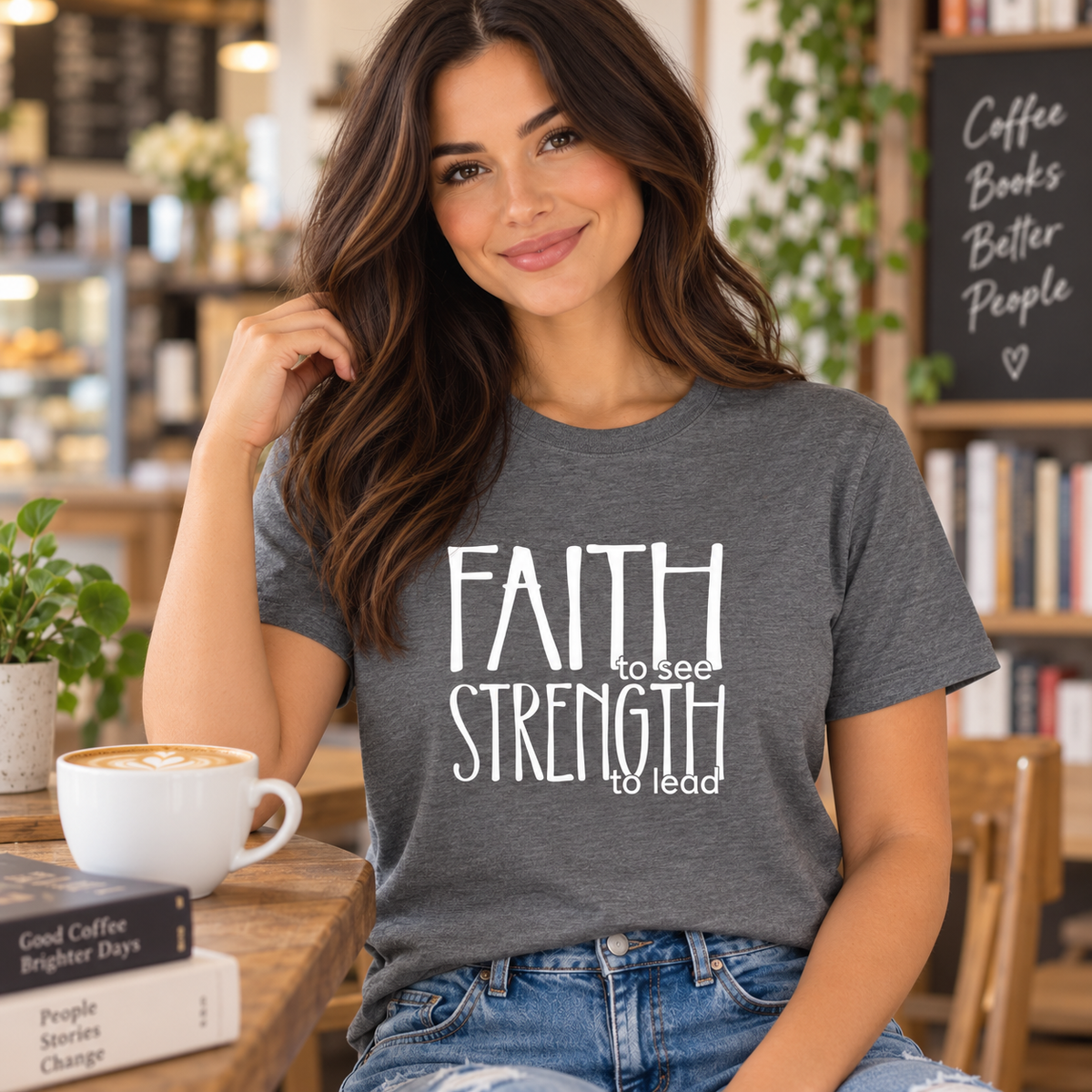 A woman wearing a heather dark gray graphic t-shirt with the white text 'FAITH to see STRENGTH to lead' sits in a cozy coffee shop environment.