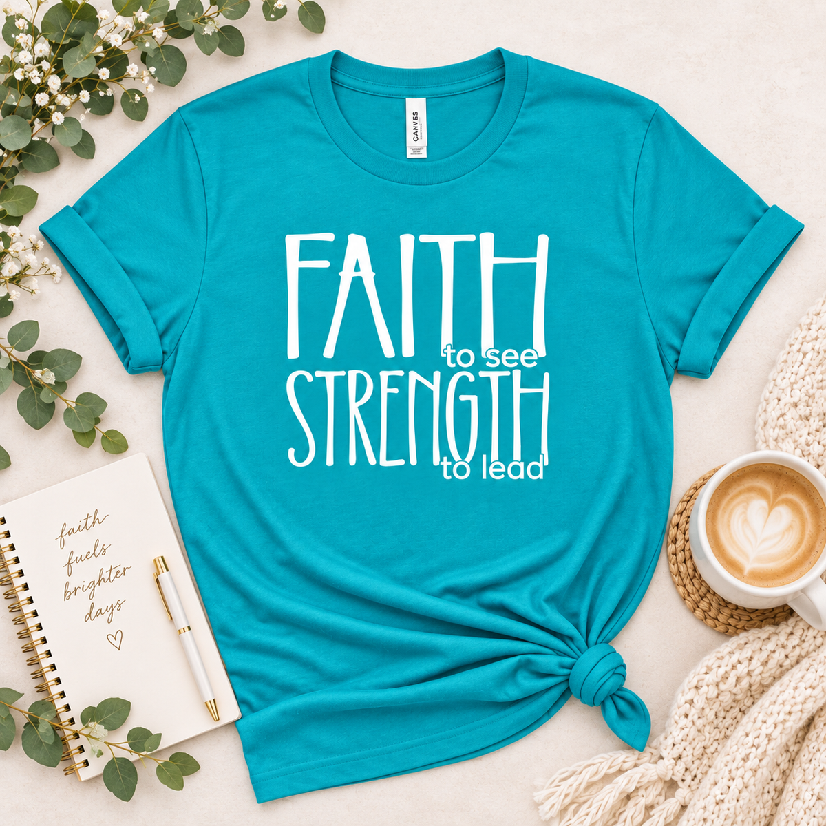 A turquoise short-sleeve T-shirt laid flat on a light beige surface, styled with eucalyptus leaves, a notebook, a pen, a coffee cup, and a knitted blanket. The shirt has white text reading 'FAITH to see STRENGTH to lead' and is tied in a knot at the lower right hem.