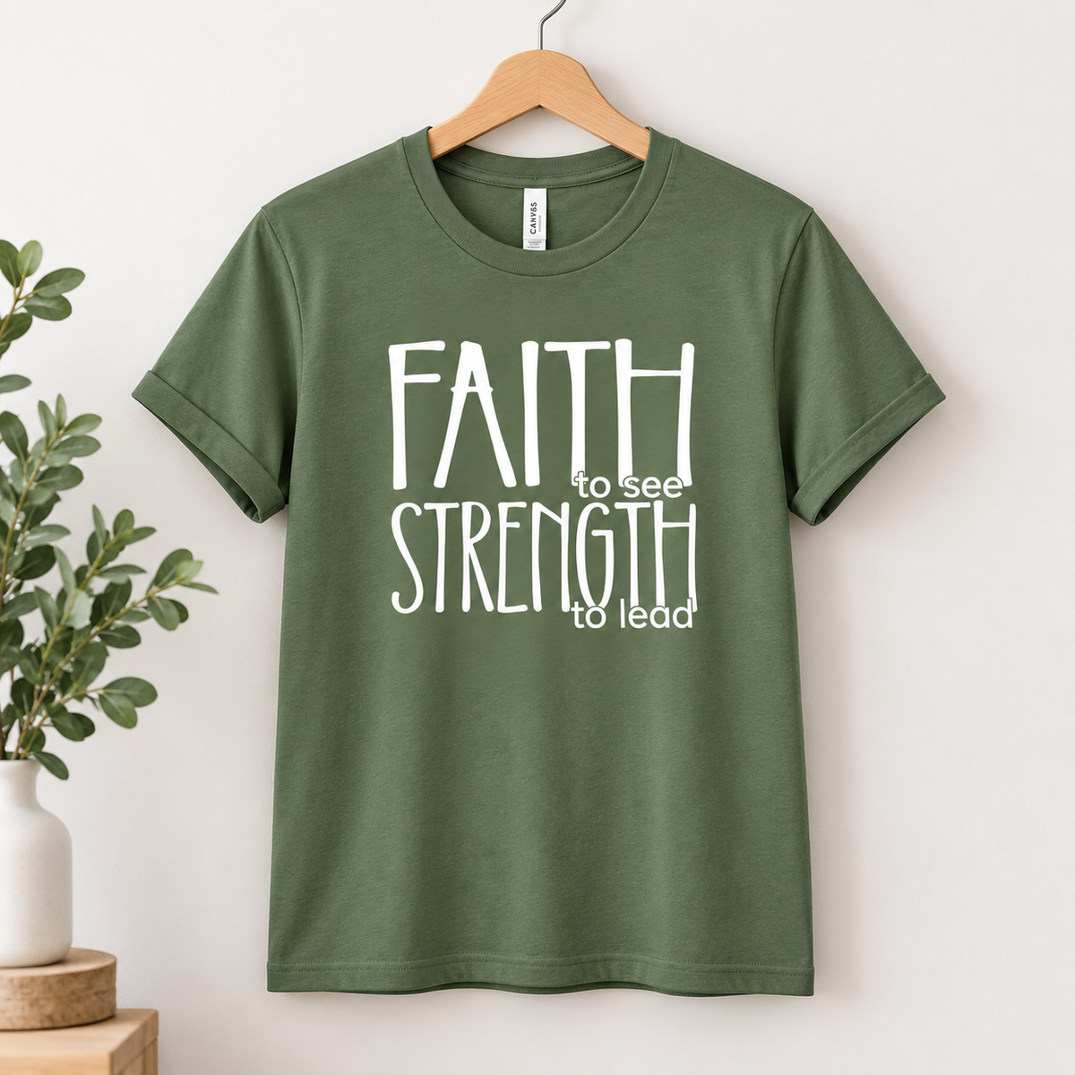 An olive green short-sleeve t-shirt hanging on a wooden hanger against a light beige wall, featuring the white text 'FAITH to see STRENGTH to lead' printed on the front. A small white plant pot is visible on the left side.
