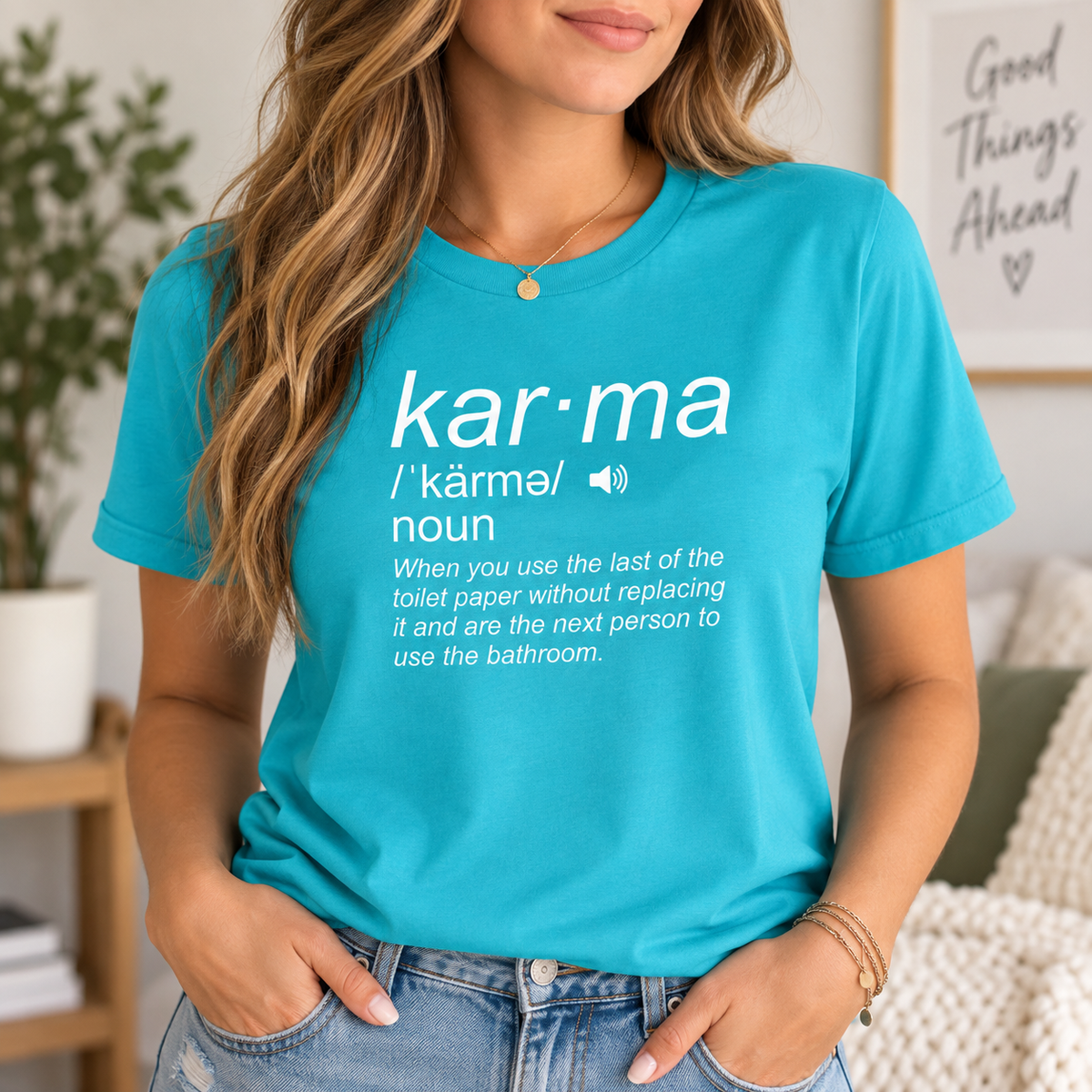 A woman wearing a bright turquoise blue graphic t-shirt with a white dictionary-style definition of the word 'kar-ma' printed on the front. The setting is a cozy indoor room with a framed print on the wall and a plant in the background.
