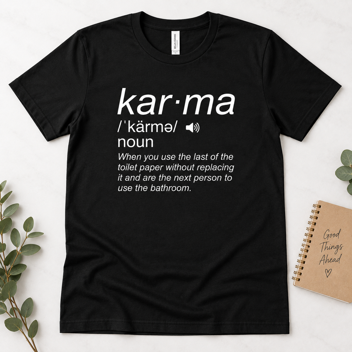 A black short-sleeve graphic T-shirt laid flat on a light surface, with white printed text defining the word 'kar·ma' and a humorous bathroom-related definition. A small notebook and greenery are placed beside it as styled props.