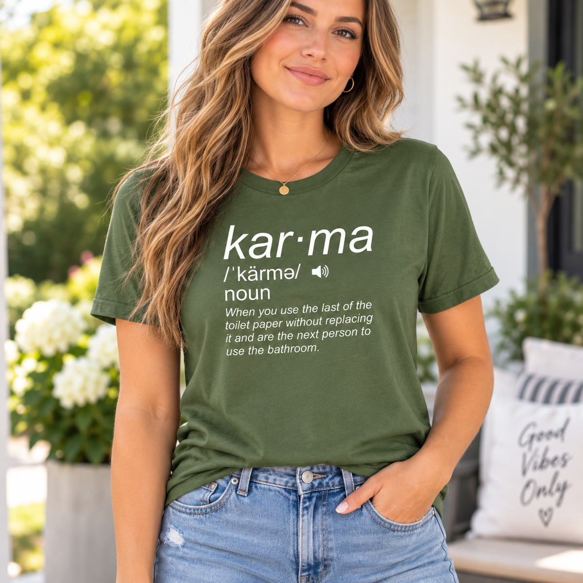 A woman wearing an olive green graphic t-shirt with a white dictionary-style definition print for 'kar-ma'. The background is a softly blurred outdoor patio setting with green foliage and a white cushion.