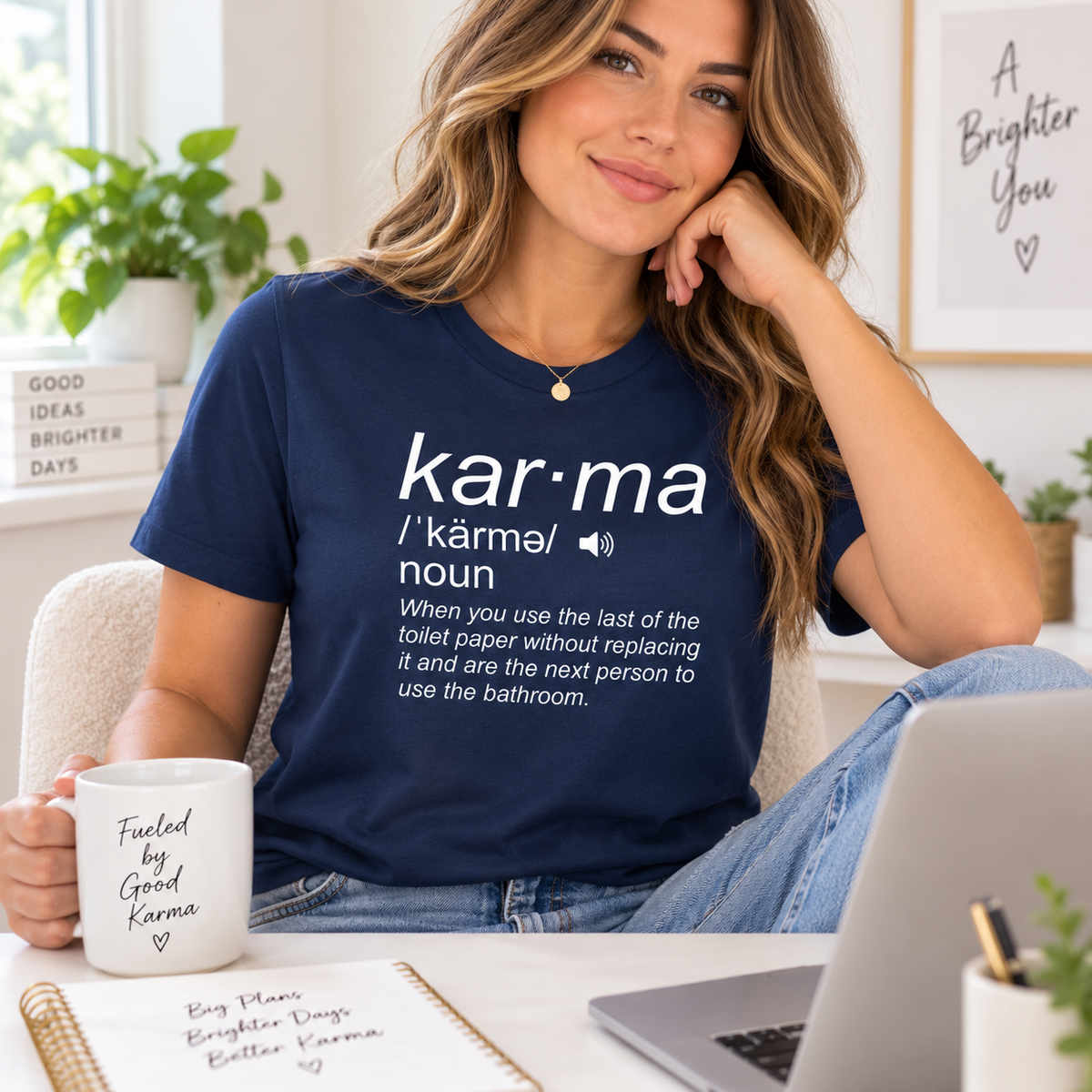 A woman sitting in a bright room wearing a navy blue graphic T-shirt with a white dictionary-style definition of 'kar·ma'. She is holding a white mug that reads 'Fueled by Good Karma' and has a laptop in front of her.