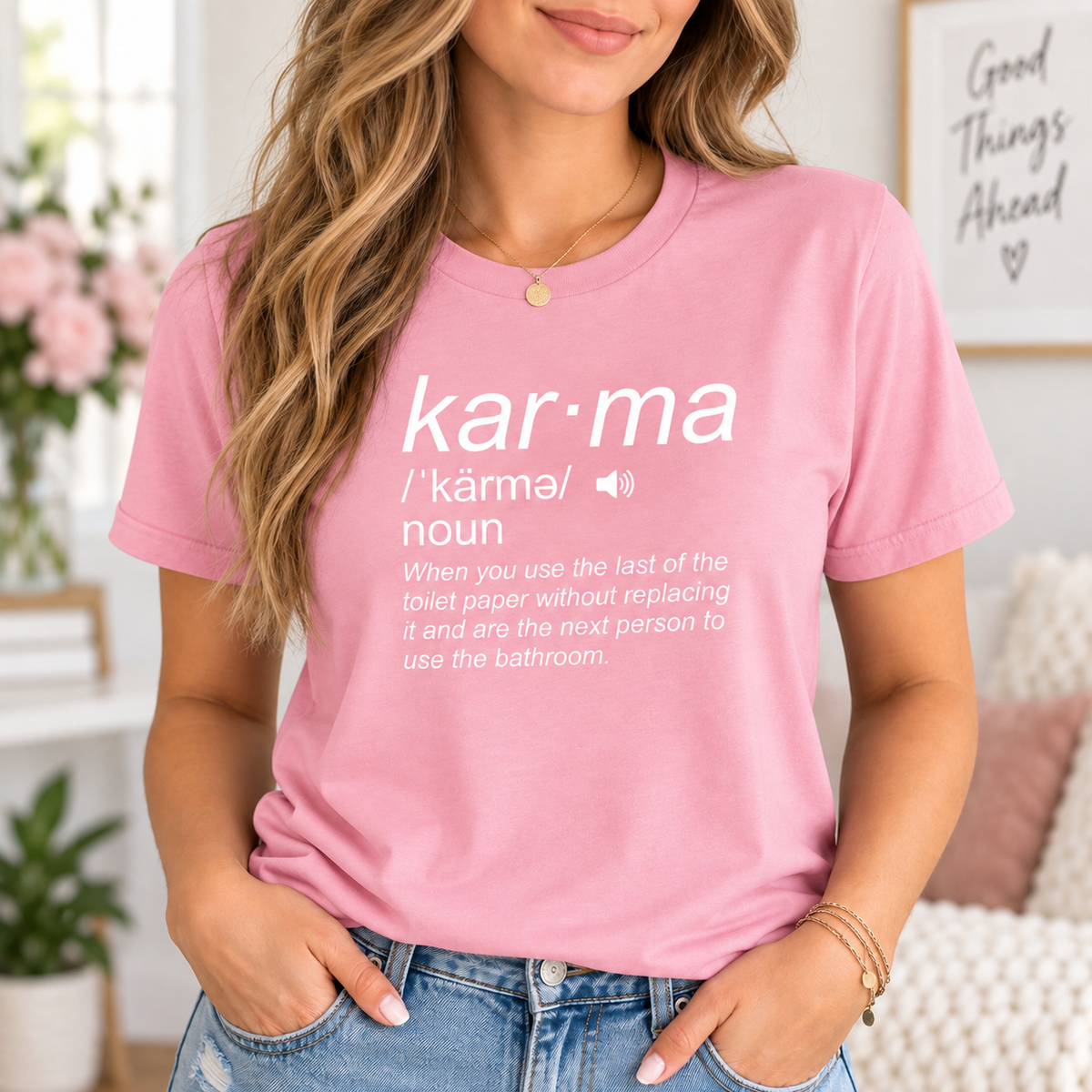 A woman wearing a pink graphic t-shirt with a white dictionary-style definition of the word 'kar-ma' printed on the front, paired with blue jeans in a brightly lit indoor setting.