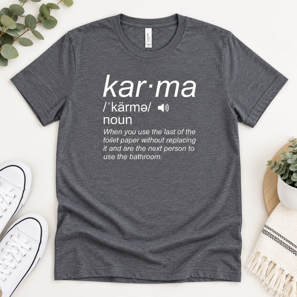 A dark heather gray graphic t-shirt laid flat on a light beige background, styled with white sneakers, a small potted plant, and a woven textile. The shirt features a white printed dictionary-style definition of the word 'kar·ma'.